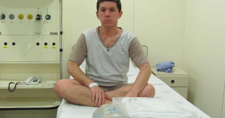 Imagem de Marcelo sentado na cama hospitalar, de pernas cruzadas, vestido camiseta e short de pijama.