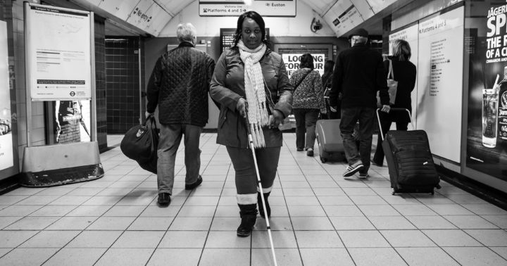 Imagem de uma mulher negra segurando a bengala-guia andando numa estação de metrô de Londres com várias pessoas ao redor.