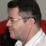 Imagem de David com rosto sorridente, visto de perfil. O cabelo anteriormente raspado, já voltou a crescer. Ele usa óculos de grau e a parte externa do IC está ligada por um fio ao computador, para fazer a programação necessária.