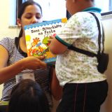 Imagem da fonoaudiologa Lilian segurando um livro infantil de capa colorida, que não dá pra enxergar direito, na foto. Um menino, implantado, olha para a capa do livro.