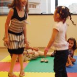 Imagem de três meninas entre 6 e 10 anos. Duas estão em pé, pulando de frente uma pra outra e uma terceira aparece sentada no tapete de E.V.A. colorido.