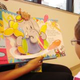 Imagem da fono Lilian segurando o livro infantil, que mostra um desenho de elegante, feito em dobradura pra um menino pequeno, que além do implante coclear, usa óculos de grau também.