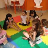 Varias crianças aparecem sentadas no tapete de e.v.a. colorido, enquanto aguardam a próxima atividade. Todas utilizam o implante coclear.