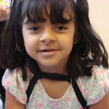 A menina Duda, de 7 anos, usuária do implante coclear, aparece olhando para a câmera com um sorriso discreto. Ela é pequena, magra e tem cabelo e olhos castanhos bem expressivos.