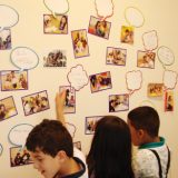 As crianças olham para uma parede repleta de fotos delas e alguns balões onde há alguns nomes escritos.