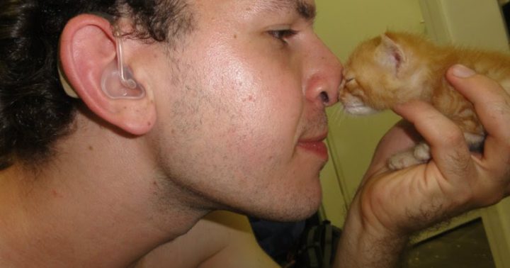 Imagem de Guilherme, de perfil, de modo que aparece o aparelho auditivo que ele usa na orelha não implantada. Ele segura um filhote de gato, que também aparece de perfil. Guilherme o segura com apenas uma mão, de modo que seu nariz e o do gato, se tocam. O gato está de olhinhos fechados.