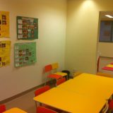 Imagem da sala onde as crianças ficam. Duas mesas quadradas, amarelas, baixas são acompanhadas de cadeiras pequenas amarelas e laranjas. Na parede, há várias cartolinas verdes e amarelas com calendários de atividades das crianças.