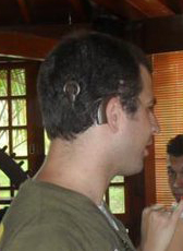 Foto do Marcus visto de perfil, com a parte externa do implante coclear aparecendo. Uma parte do aparelho fica atrás da orelha, ligada por um fio (chamado de antena) até outra parte que fica colada no crânio, por meio de imã.