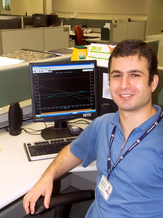 Foto de um homem jovem com um crachá no pescoço, sentado em frente a um computador em ambiente de trabalho, olhando para frente e sorrindo.