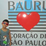 Kadu de óculos escuros de aro vermelho, em frente a um painel, onde está escrito: Bauro, coração de São Paulo.