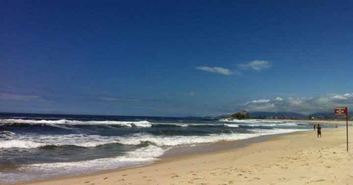 Imagem da praia de Itauna, em Saquarema. O céu azul com poucas nuvens, o mar num tom azul profundo, revolto, e a areia bege clara. Há também uma placa vermelha indicando sinal de perigo.
