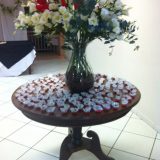 Imagem de uma mesa de madeira, redonda, com vários docinhos em forminhas vermelhas e brancas, sobre ela. Há também um grande arranjo de flores no centro da mesa.