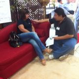 O jornalista da mídia local (Londrina PR) conversa com uma das participantes do evento. Ambos estão sentados num sofá vermelho