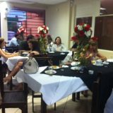 Imagem do espaço onde foi o evento. Algumas mesas com toalha preta e branca, com arranjos de rosas vermelhas e brancas. Ao fundo, algumas pessoas sentadas nas mesas.