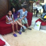 Imagem de 3 crianças, duas meninas e um menino, sentados num sofá vermelho.