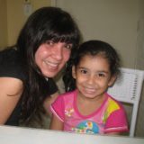 Foto de Mônica, sorrindo, ao lado da pequena Milena, que também sorri para a câmera.