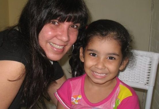 Foto de Mônica, sorrindo, ao lado da pequena Milena, que também sorri para a câmera.